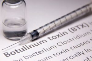 A glass medical vial containing a clear liquid next to an empty syringe and needle sitting on a table surface. There is text on the table surface describing botulinum toxin.