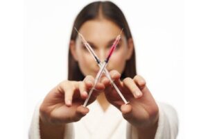 A woman with her face blurred out holding two medical needles in front of her, the needles are crisscrossed. One needle is full of red liquid, the other needle is empty.